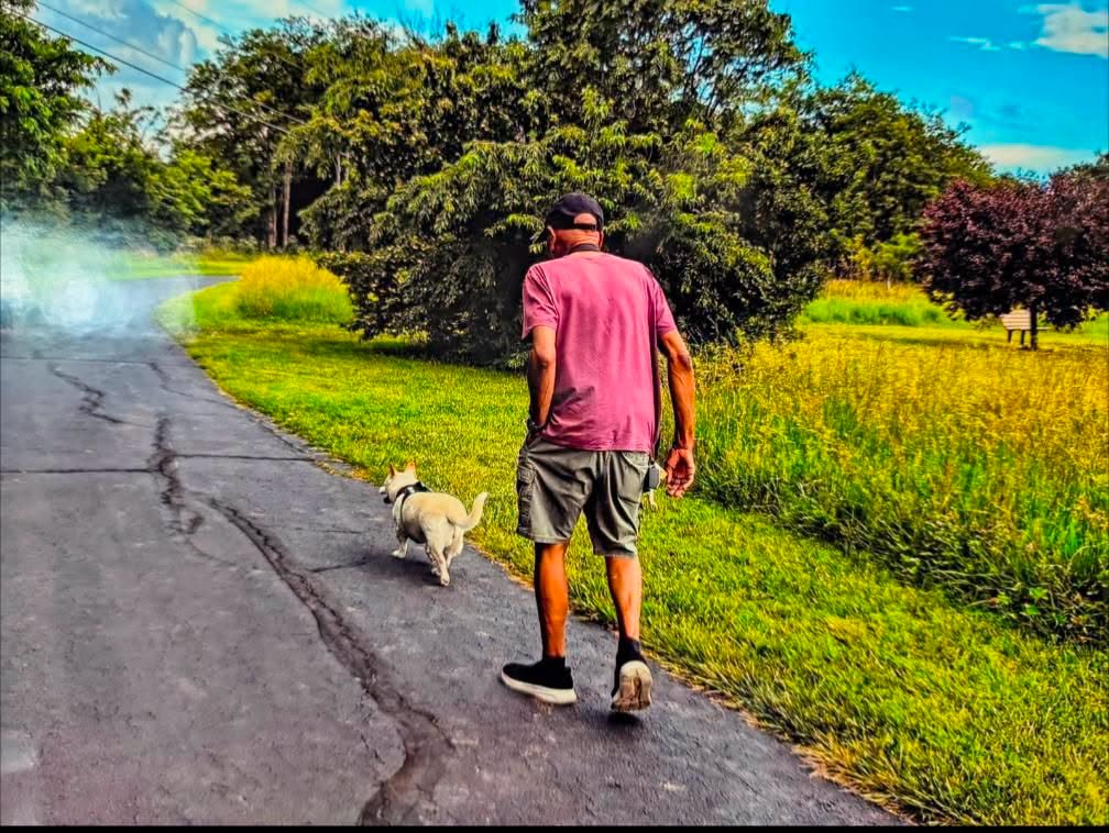 Jim walking down a tree-lined path on a sunny day, Mimi trotting ahead of him on her own four legs