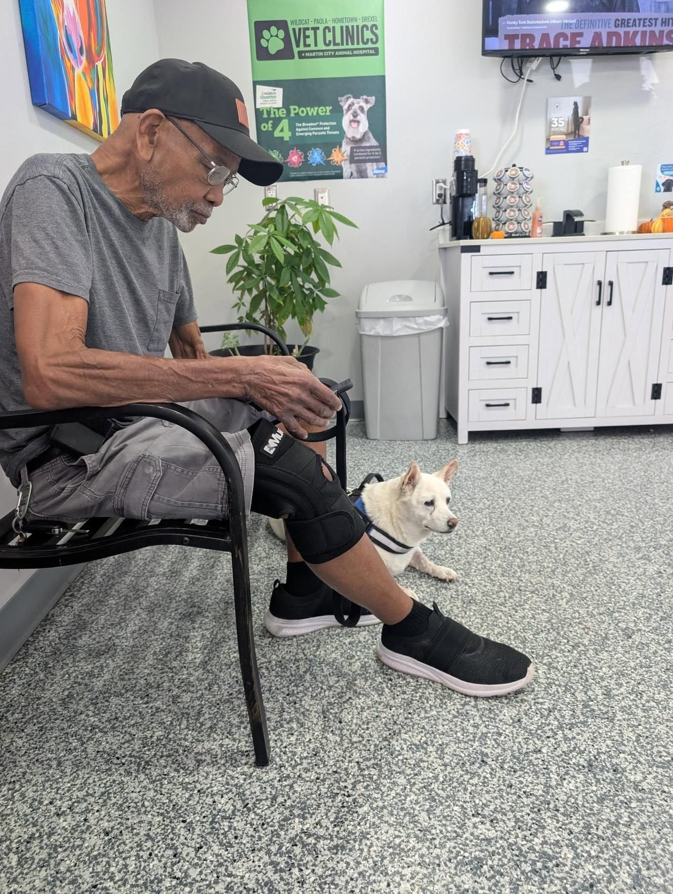Jim, in a black ball cap and knee brace, sitting in a vet clinic waiting room with Mimi lying calmly at his feet